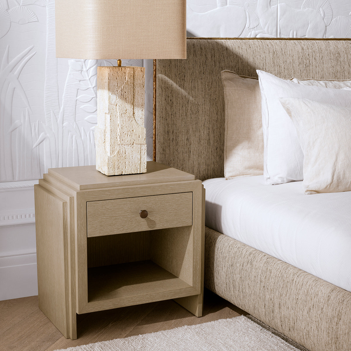 A bedroom vignette featuring the Eichholtz Nightstand ArtDeco — geometric brass inlay, warm tones, and a sculpted base beside a styled upholstered bed.