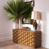 Dresser Rubicon — geometric carved oak dresser styled with a tropical palm, antique mirror, and table lamp in a white-panelled room with herringbone floors.
