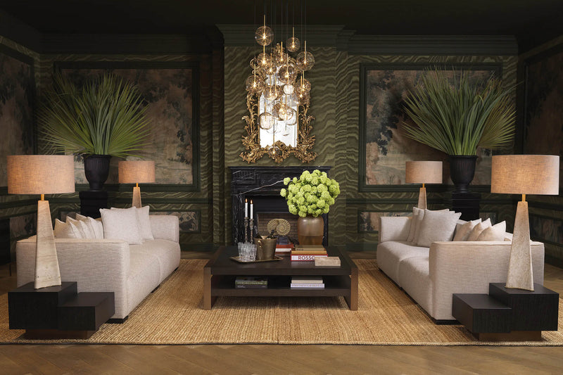 A dramatic Eichholtz living-room vignette — cream sofas facing each other across a low stone-topped coffee table, crystal chandelier overhead