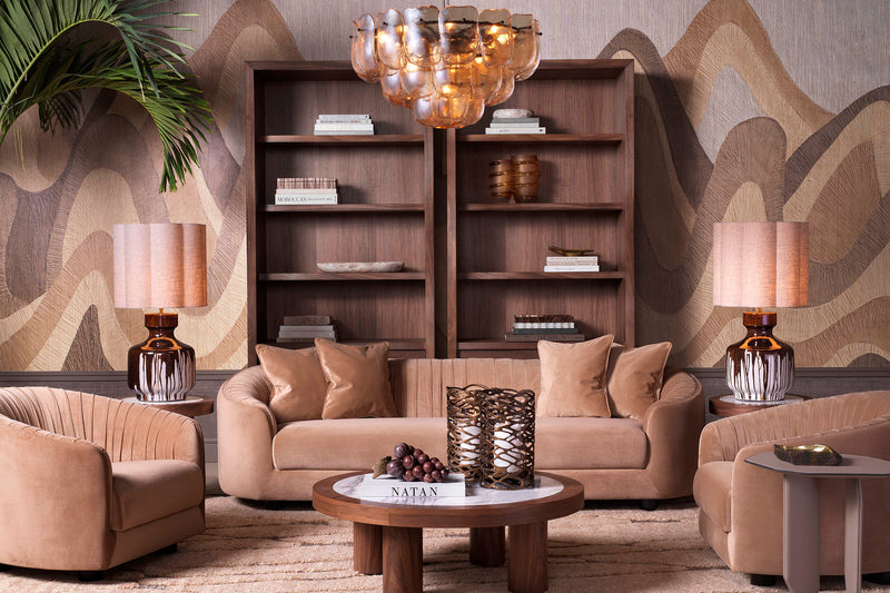 A curated Eichholtz living-room vignette — a tan velvet sofa flanked by twin lamps, set against walnut bookcases and an amber cluster chandelier