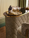Close up of a curved upholstered bar with patterned fabric, brass trim, crystal cocktail glasses, and a decorative tray styled in a luxury interior setting.