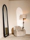 Sculptural accent chair paired with a tall black framed floor mirror and table lamp, styled against textured neutral walls in a refined interior setting.