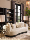 Curved ivory sofa with textured accent pillows, black shelving, table lamps, and layered neutral décor in an elegant living room setting.