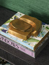 Close up of sculptural brass decorative object displayed on stacked design books atop a dark wood surface against a green backdrop.