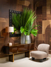 Luxury console table with rich wood finish, tall green foliage in textured vases, accent chair, and table lamp set against geometric wood wall panels.