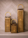 Set of three brass lanterns with woven panels and loop handles, softly illuminated and arranged against a light stone wall.