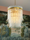 Sculptural glass chandelier with layered feather like elements and brass accents suspended against a painted mural backdrop.