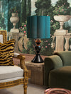 Decorative table lamp with teal shade on stone side table beside green sofa and carved accent chair, styled against a painted mural wall.
