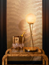 Brass table lamp with glowing glass globe styled on a wood side table with champagne bucket and glassware against a textured wall.